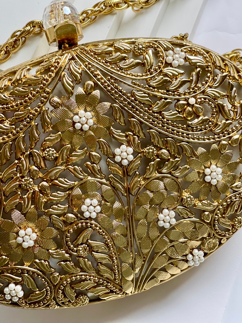 May include: A gold metal clutch bag with a floral design and white pearl accents. The bag has a chain strap.