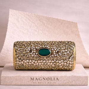 Könnte beinhalten: Eine rechteckige, goldfarbene Clutch, verziert mit zahlreichen klaren, funkelnden Steinen und kleinen weißen Perlen. Die Clutch hat einen zentralen, oval geformten, dunkelgrünen Stein. Der Markenname "MAGNOLIA BY NAJLA MALIK" ist darunter aufgedruckt.