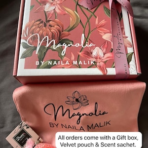 May include: A gift set with a floral-patterned box, a velvet pouch, and a scent sachet. The box is adorned with the word "Magnolia" and the brand name "NAILA MALIK". A pink ribbon is tied around the box. Text on the box reads "All orders come with a Gift box, Velvet pouch & Scent sachet."