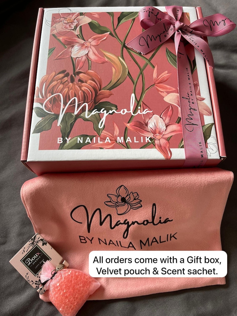 May include: A gift set with a floral-patterned box, a velvet pouch, and a scent sachet. The box is adorned with the word "Magnolia" and the brand name "NAILA MALIK". A pink ribbon is tied around the box. Text on the box reads "All orders come with a Gift box, Velvet pouch & Scent sachet."