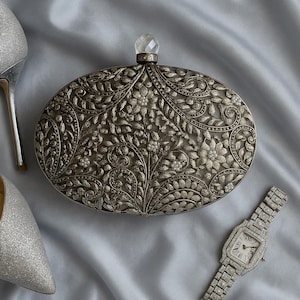 Puede incluir: Bolso de mano plateado, de forma ovalada, con intrincados diseños florales y de hojas. Una gema transparente facetada corona el bolso. Un reloj plateado con esfera rectangular y un zapato de tacón plateado también están en la imagen.