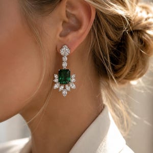May include: A drop earring featuring a large, rectangular emerald-green center stone surrounded by a cluster of clear, marquise-cut stones. The top section consists of a smaller, clear, pear-shaped stone and a cluster of tiny, clear, round stones.