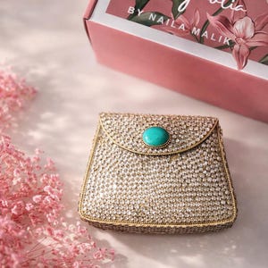 Puede incluir: Un pequeño bolso de mano cuadrado, de color dorado, cubierto de cristales brillantes, con un cierre de piedra turquesa. El bolso de mano está sobre un fondo rosa suave, con una caja floral rosa en el fondo. La caja dice "Magnolia BY NAILA MALIK".