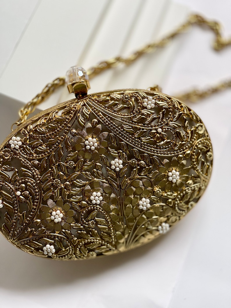 May include: A gold metal clutch bag with an intricate floral design and white pearl accents. The bag has a chain strap.