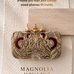 May include: A rectangular clutch bag featuring intricate gold and deep red floral embroidery on a grey base, with a gold metal clasp. Text reads: iPhone pro max and similar sized phones fit inside. MAGNOLIA BY NAILA MALIK.
