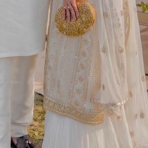 May include: A white embroidered dress with a gold clutch. The dress has a sheer overlay and a pleated skirt.