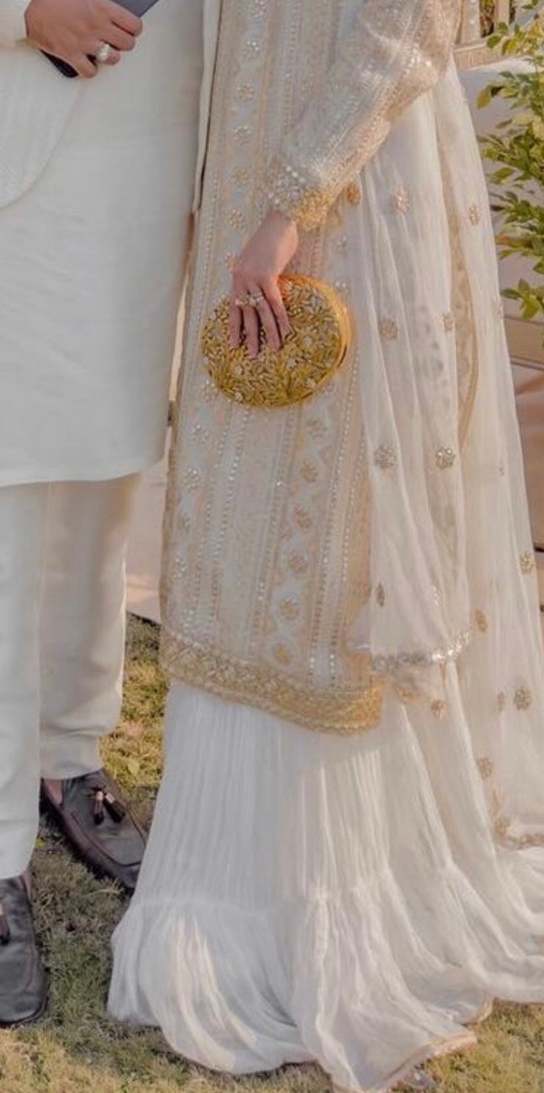 May include: A white embroidered dress with a gold clutch. The dress has a sheer overlay and a pleated skirt.
