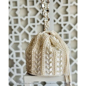 May include: Cream-coloured beaded handbag with a drawstring closure. The bag is adorned with pearls and rhinestones, featuring a pearl-studded handle and decorative accents. The bag is on a white surface.