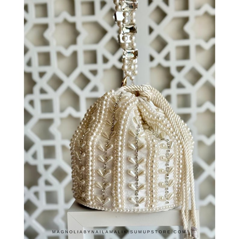 May include: Cream-coloured beaded handbag with a drawstring closure. The bag is adorned with pearls and rhinestones, featuring a pearl-studded handle and decorative accents. The bag is on a white surface.
