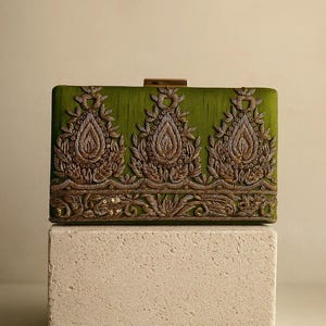 Puede incluir: Clutch rectangular verde oliva con bordados dorados ornamentados. Presenta tres diseños bordados centrales en forma de lágrima y un cierre dorado. Se exhibe sobre un bloque texturizado blanquecino, resaltando los detalles.