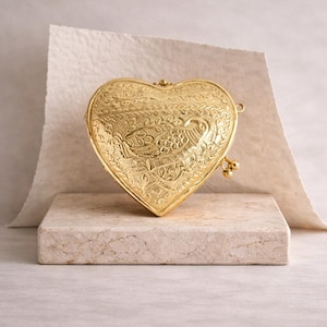 Può includere: Una pochette dorata a forma di cuore con intricati disegni floreali e di uccelli in rilievo. La pochette è appoggiata su un blocco di pietra rettangolare, con uno sfondo neutro. La chiusura e le piccole perle decorative sono visibili.