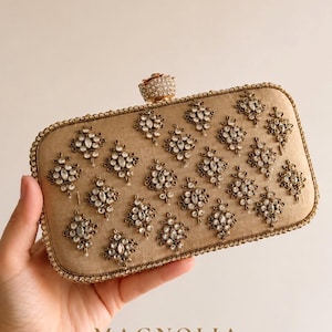 Peut inclure: Une pochette rectangulaire dorée ornée de motifs floraux complexes. La pochette est ornée de pierres précieuses claires et étincelantes et d'un fermoir décoratif. La marque "MAGNOLIA" est imprimée en bas.