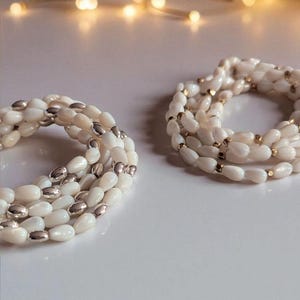 May include: Two coiled necklaces with white, oval-shaped beads and silver-colored accents. One necklace has silver accents, and the other has gold-colored accents. The necklaces are displayed on a white surface.