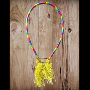 Puede incluir: Un collar de cuerda arcoíris con un colgante de borla amarilla. El collar presenta una cuerda trenzada en tonos del arcoíris. El colgante es una cascada de hebras amarillas rizadas, suspendidas de un cierre plateado.