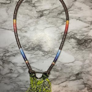 May include: A unique necklace featuring a brown and black patterned cord with colorful accents of yellow, red, and blue. The necklace has a silver-colored clasp and a fringe of silver and neon green chain.