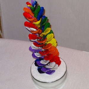May include: A rainbow-colored pine cone ornament with each scale painted a different color, sitting on a bed of white glitter in a clear glass jar.