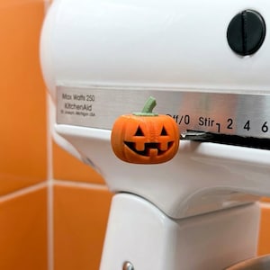 Puede incluir: Una cubierta de perilla con forma de calabaza jack-o'-lantern naranja unida a la palanca de control de velocidad de una batidora de pie KitchenAid blanca. La batidora tiene un panel de control plateado con el texto Max Watts 250 KitchenAid.