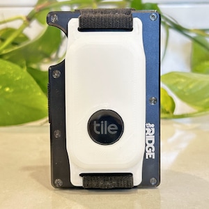 May include: A black and blue Ridge wallet with a white Tile tracker attached. The Tile tracker has a black circle with the word "tile" in white. The wallet has a black elastic band and is on a light-coloured surface.