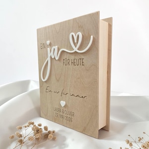 Könnte beinhalten: Eine hellfarbene Holzbox mit Klappdeckel, verziert mit dem weißen 3D-Schriftzug Ein ja FÜR HEUTE und einem Herz. Darunter ist der Text Ein wir für immer. eingraviert, gefolgt von einem kleinen weißen Herz, LAURA & OLIVER und 23. MAI 2026.