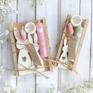 Könnte beinhalten: Zwei Holztabletts mit Osterdekorationen. Jedes Tablett enthält einen weißen Hasen-Ausschnitt, eine Kerze, eine kleine Keramikschale und getrocknete rosa Blumen. Die Kerzen sind rosa und grün, und die Hasen haben Herz-Ausschnitte.