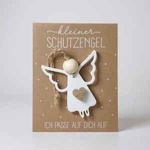 May include: A white wooden angel ornament with a wooden heart detail, attached to a brown card with the German text "kleiner SCHUTZENGEL" and "ICH PASSE AUF DICH AUF". The angel has a wooden bead head and twine for hanging. The card has white dot details.