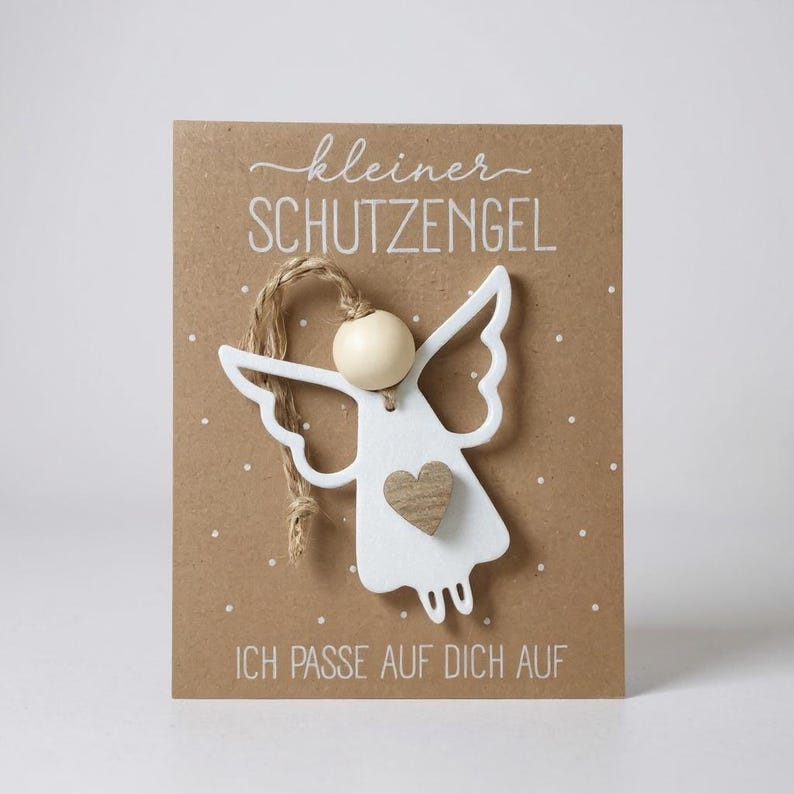 May include: A white wooden angel ornament with a wooden heart detail, attached to a brown card with the German text "kleiner SCHUTZENGEL" and "ICH PASSE AUF DICH AUF". The angel has a wooden bead head and twine for hanging. The card has white dot details.