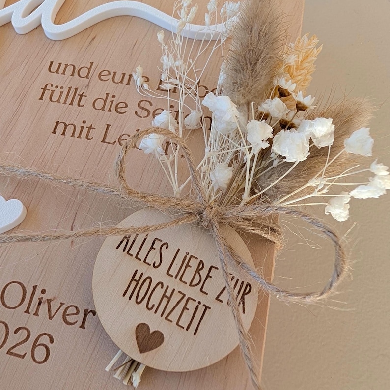 May include: A light-colored wooden plaque featuring engraved text including "Oliver" and "2026," topped with a white script cutout. A circular wooden tag tied with twine reads "ALLES LIEBE ZUR HOCHZEIT" with a heart icon, accented by dried white flowers.