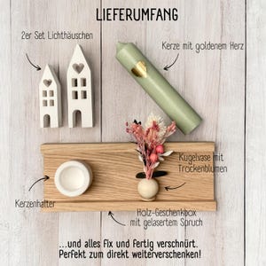 Peut inclure: Une bo&icirc;te cadeau en bois contenant une bougie, deux petites maisons, un vase avec des fleurs s&eacute;ch&eacute;es et un bougeoir. La bo&icirc;te est &eacute;tiquet&eacute;e "LIEFERUMFANG" et comprend le texte "... und alles fix und fertig verschn&uuml;rt. Perfekt zum direkt weiterverschenken!"