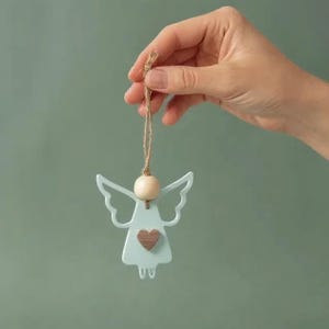 May include: A light blue angel ornament with a wooden heart and bead, suspended by a natural twine string. The angel has detailed wings and a simple, elegant design. The ornament is held against a muted green background.