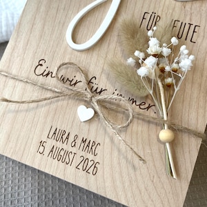 May include: A light-colored wooden plaque featuring the German text "Ein wir f&uuml;r immer" and "LAURA & MARC 15. AUGUST 2026" in black lettering. A jute twine bow with a small white wooden heart and a dried floral arrangement are attached to the front.