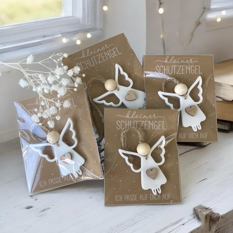 May include: Four small, white angel ornaments with wooden bead heads and heart-shaped accents are displayed on brown cards. The cards have the text "kleiner SCHUTZENGEL" and "ICH PASSE AUF DICH AUF". The angels are packaged in clear plastic.