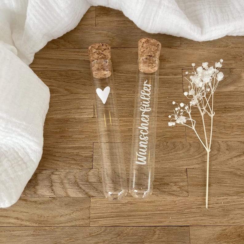 May include: Two clear glass test tubes with cork stoppers. One tube features a white heart graphic, and the other tube features the white text Wunscherf&uuml;ller. A small sprig of dried white baby's breath flowers is positioned next to the glass tubes.