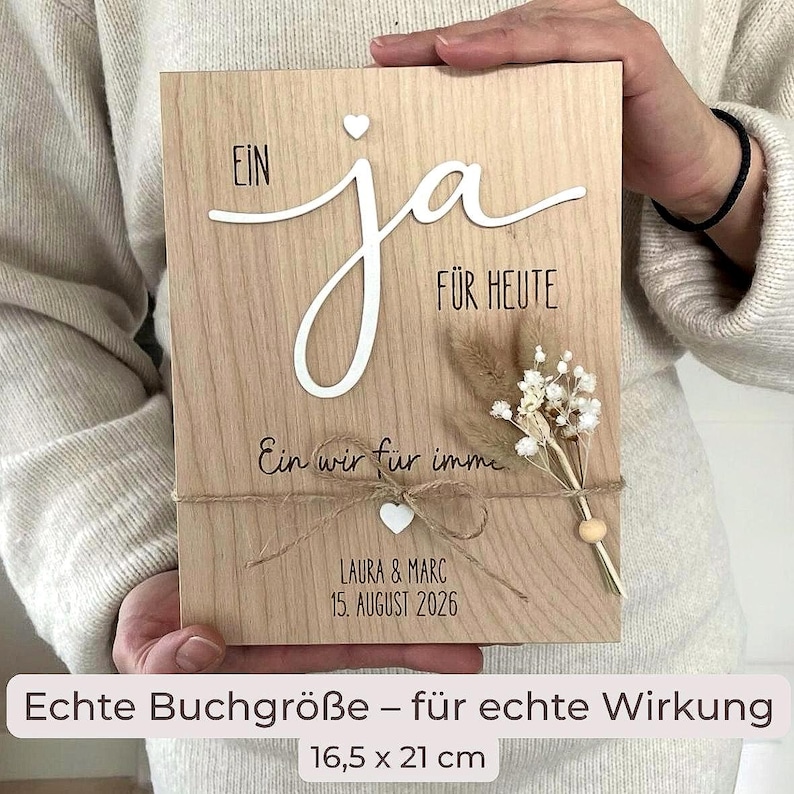May include: A rectangular light wood plaque measuring 16.5 by 21 cm, featuring the German text "EIN ja F&Uuml;R HEUTE" and "Ein wir f&uuml;r immer" with "LAURA & MARC 15. AUGUST 2026". It includes a small white heart, twine bow, and dried flowers.