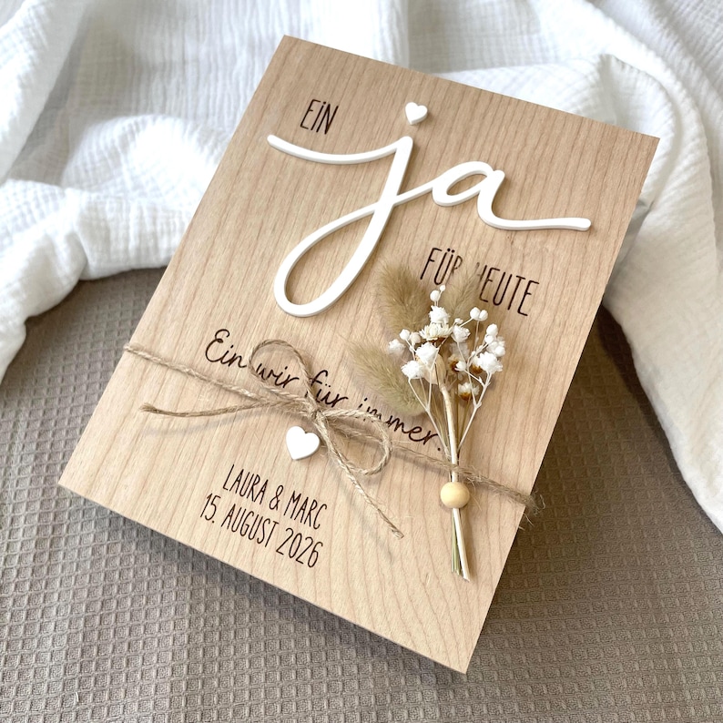 May include: A rectangular wooden plaque featuring the German text "EIN ja F&Uuml;R HEUTE" and "Ein wir f&uuml;r immer" with the names "LAURA & MARC" and the date "15. AUGUST 2026". It is decorated with a small dried flower bouquet, twine, and white hearts.