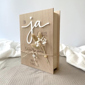 May include: A rectangular wooden box with a light wood grain finish. The front features the white 3D text "Ein ja" above the printed text "f&uuml;r heute" and "Ein wir f&uuml;r immer". Below, "Laura & Marc" and "15. August 2026" are printed. A small dried flower bouquet is tied with twine.