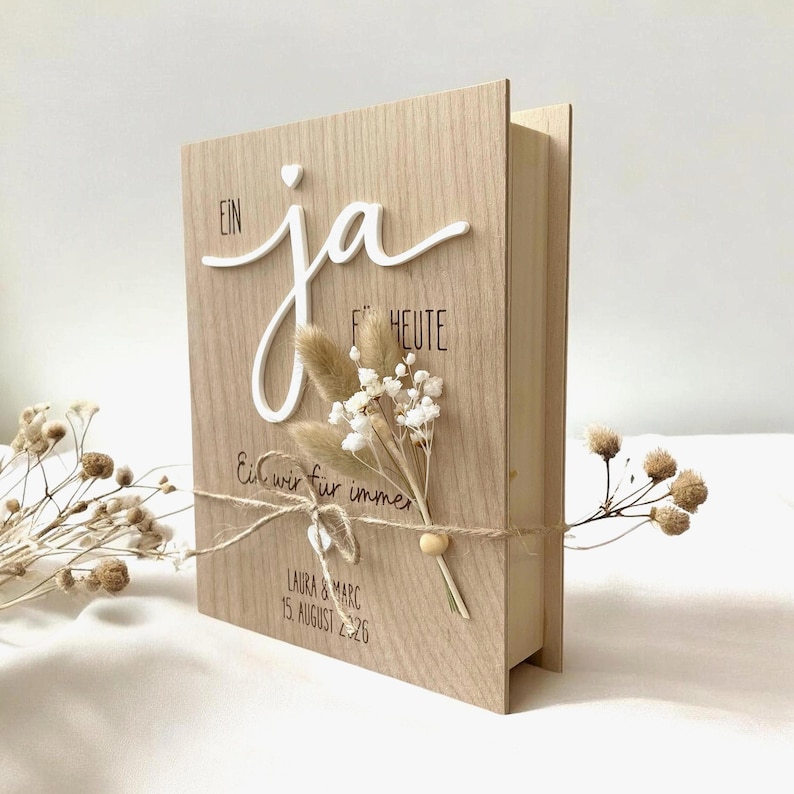 May include: A light wood rectangular box with the German text "EIN JA F&Uuml;R HEUTE" and "Ein wir f&uuml;r immer" printed on the front. It features a raised white "ja" script, a small dried flower bouquet tied with twine, and the names "LAURA & MARC 15. AUGUST 2026".