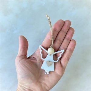 May include: A white angel ornament with a wooden heart and bead, suspended by a natural twine string. The angel has cut-out wings and a simple dress design. A charming, handcrafted decoration.