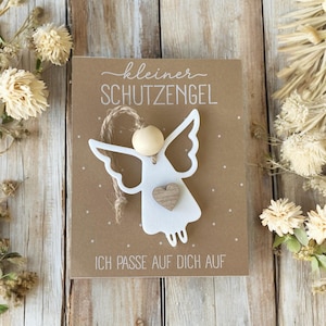May include: A white angel-shaped ornament with a wooden bead head and a small wooden heart on a brown card. The card has the text "kleiner SCHUTZENGEL" and "ICH PASSE AUF DICH AUF". The background is decorated with dried flowers.