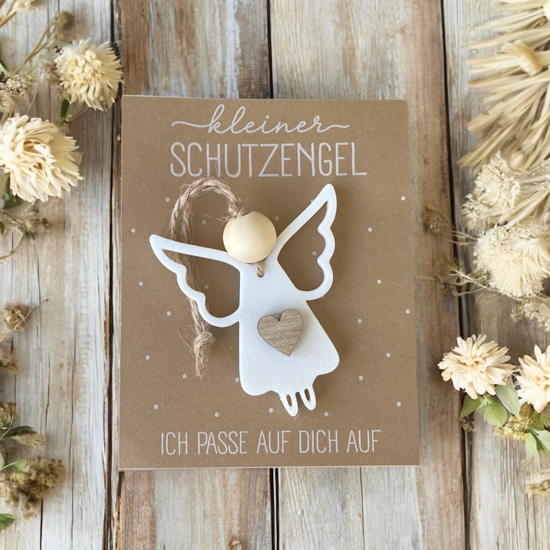 May include: A white angel-shaped ornament with a wooden bead head and a small wooden heart on a brown card. The card has the text "kleiner SCHUTZENGEL" and "ICH PASSE AUF DICH AUF". The background is decorated with dried flowers.