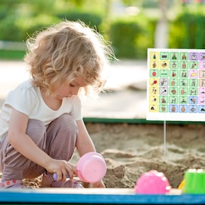 May include: A young child plays in a sandbox with pink plastic toys. A visual communication board with colorful symbols is in the background.