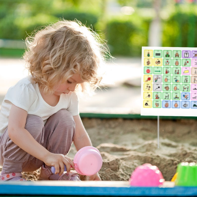 May include: A young child plays in a sandbox with pink plastic toys. A visual communication board with colorful symbols is in the background.