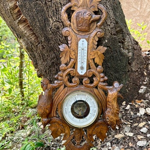 May include: A carved wooden barometer with a thermometer and a compass. The barometer has a round face with a needle that points to the current air pressure. The thermometer is a long, thin tube with a red liquid inside. The compass is a small, round dial with a needle that points to north. The barometer is decorated with carvings of birds and leaves.