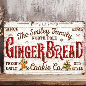 May include: A distressed wooden sign with the text "Ginger Bread Cookie Co." in red and brown. The sign includes the words "The Smiley Family North Pole" and a gingerbread man and Christmas tree graphic. The sign is on a wooden shelf.