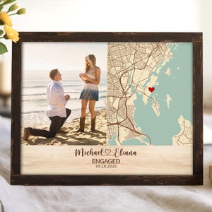 May include: A framed print featuring a photograph of a proposal on a beach, paired with a map. The map highlights a location with a red heart. The text reads "Michael & Eliana ENGAGED 09.16.2025". The frame is dark brown.