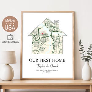 May include: A framed print with a house-shaped map design, a red heart, and the text "OUR FIRST HOME". The names "Taylor & Jacob" and an address are below the map. The print is in a wooden frame and sits on a wooden table.