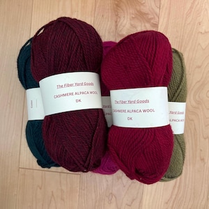 Può includere: Una collezione di gomitoli di filato colorati, tra cui tonalità bordeaux, verde acqua, rosa e verde oliva. Ogni gomitolo è avvolto con un'etichetta bianca che recita "The Fiber Yard Goods CASHMERE ALPACA WOOL DK". Il filato è disposto su una superficie di legno chiaro.