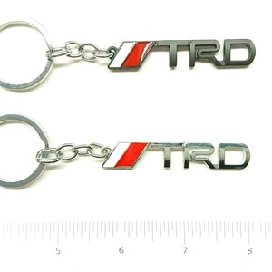 Pode incluir: Dois chaveiros prateados com uma corrente prateada e um logotipo TRD vermelho e branco.