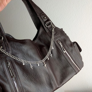 Könnte beinhalten: Eine dunkelbraune Handtasche aus Kunstleder mit einer silberfarbenen Kette, die über die Vorderseite drapiert ist. Die Kette ist mit kleinen, hängenden sternförmigen Anhängern verziert. Die Tasche hat vertikale Reißverschlusstaschen.