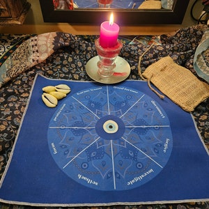 May include: A blue circular cloth with a mandala design and the words "good fortune", "disconnect", "forgive", "investigate", "self-love", "setback", "focus", "protection", "karmic lesson" around the edge. A blue evil eye charm is in the centre of the mandala. There are three seashells on the cloth.