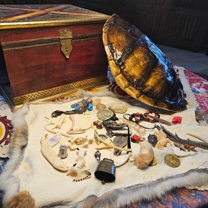Könnte beinhalten: Eine Holzkiste mit einem Messingverschluss, eine Schildkrötenschale und eine Sammlung von Knochen, Perlen und anderen Kleinigkeiten auf einer weißen Fellfläche.
